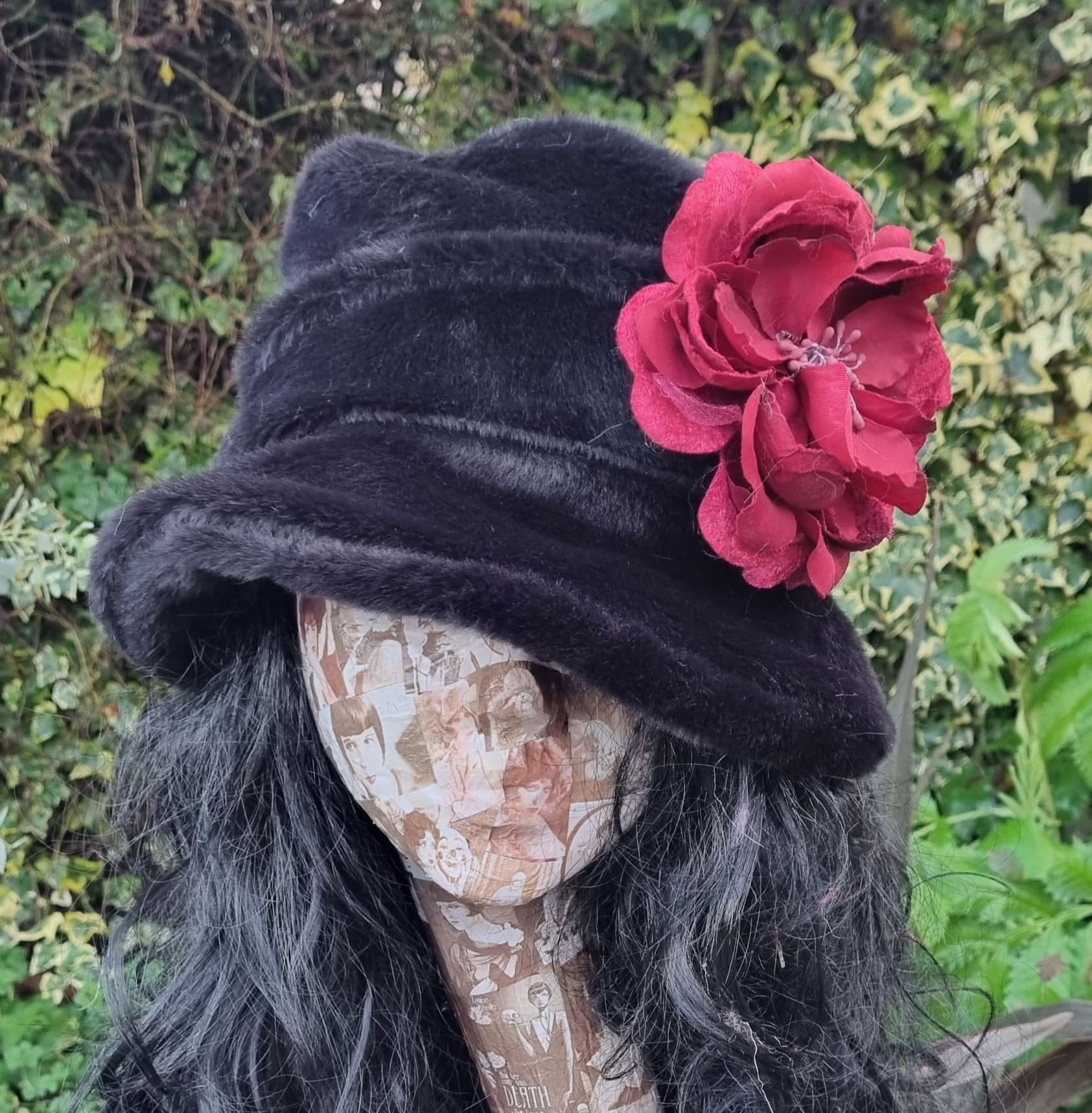 Black velvet hat with a red flower on a mannequin head against a green leafy background