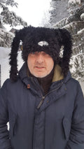 Person wearing a dark blue winter coat and a black furry hat with a logo in a snowy forest.