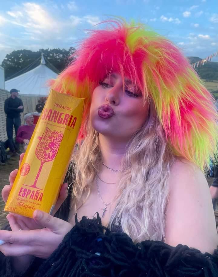 Person with colorful yellow and pink hat holding a can of 'Banga!' at an outdoor event.