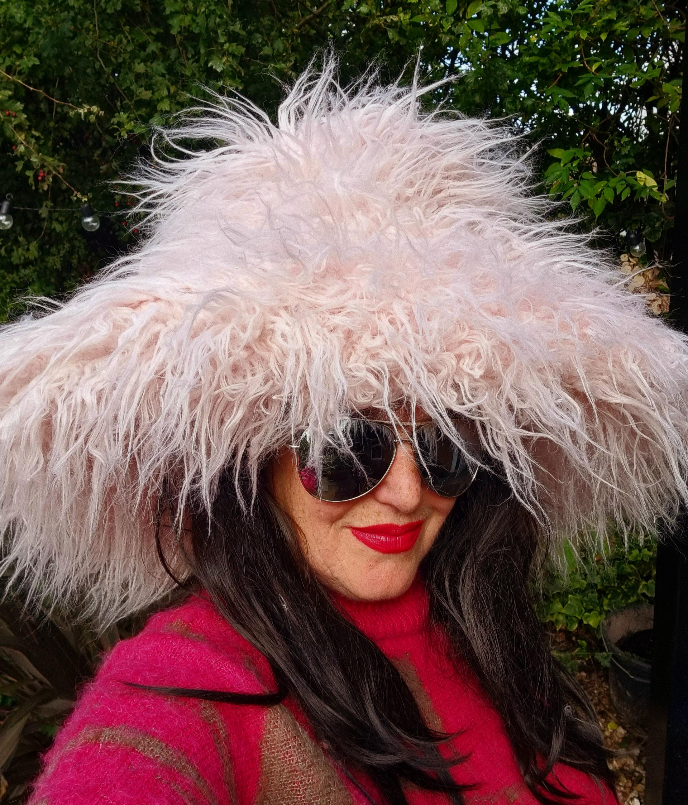 Person wearing a fluffy pink hooded coat with sunglasses and red lipstick outdoors.
